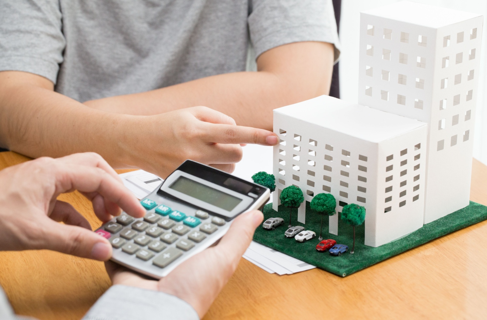 Close-up of a man sitting at a desk and holding a calculator while talking to a client who is pointing out on a miniature condominium building