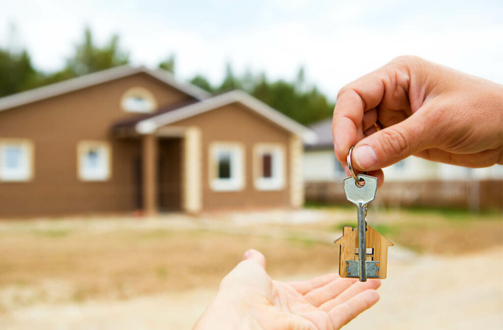 A close-up of hands exchanging an old-fashioned key on a house-shaped wooden keyring with a home blurry in the distance