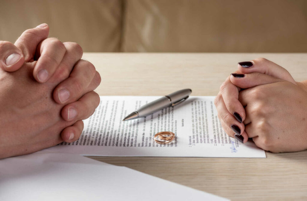 A close-up of a couple's hands as they sit across from each other with divorce papers, a pen, & gold bands on the table