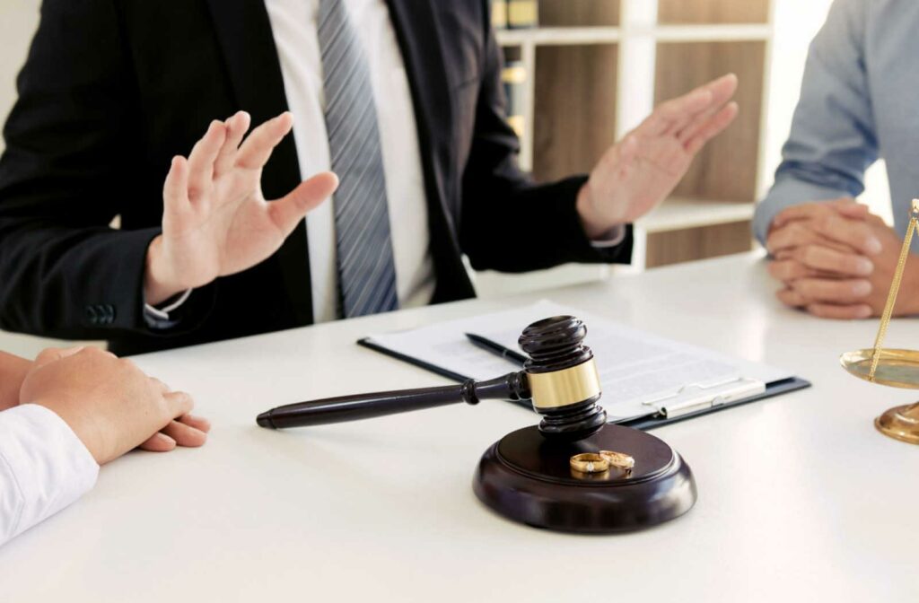 A male lawyer in a black colored suit is mediating between a man in a light blue coloured long-sleeve shirt and a woman in a white long-sleeve shirt.  A  gavel and wedding rings are on the table.