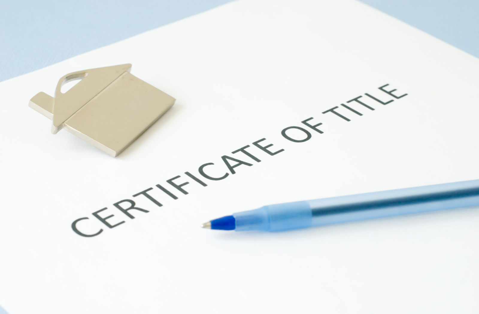 A close up of a piece of paper that says "certificate of title" on a desk with a pen and a small figure of a house.